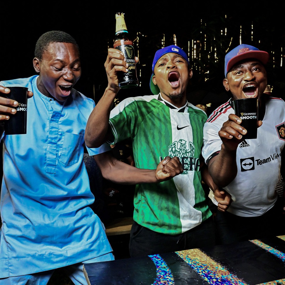 Three friends in football shirts cheer and punch the air, drinks in hand.