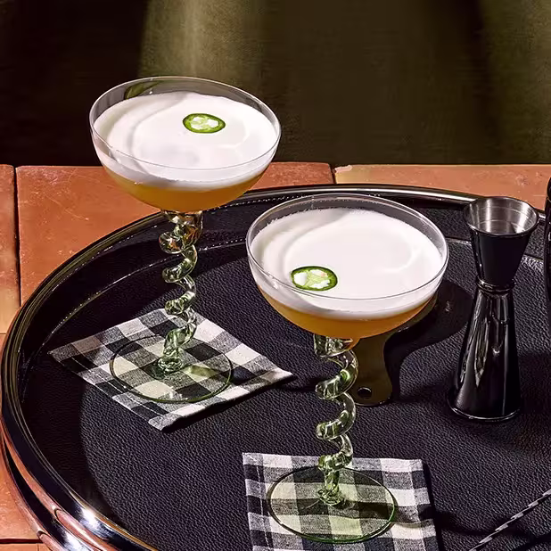 Two cocktails with frothy tops and jalapeño slices garnish, served in spiral-stem glasses on checkered napkins, placed on a tray with a metal jigger.