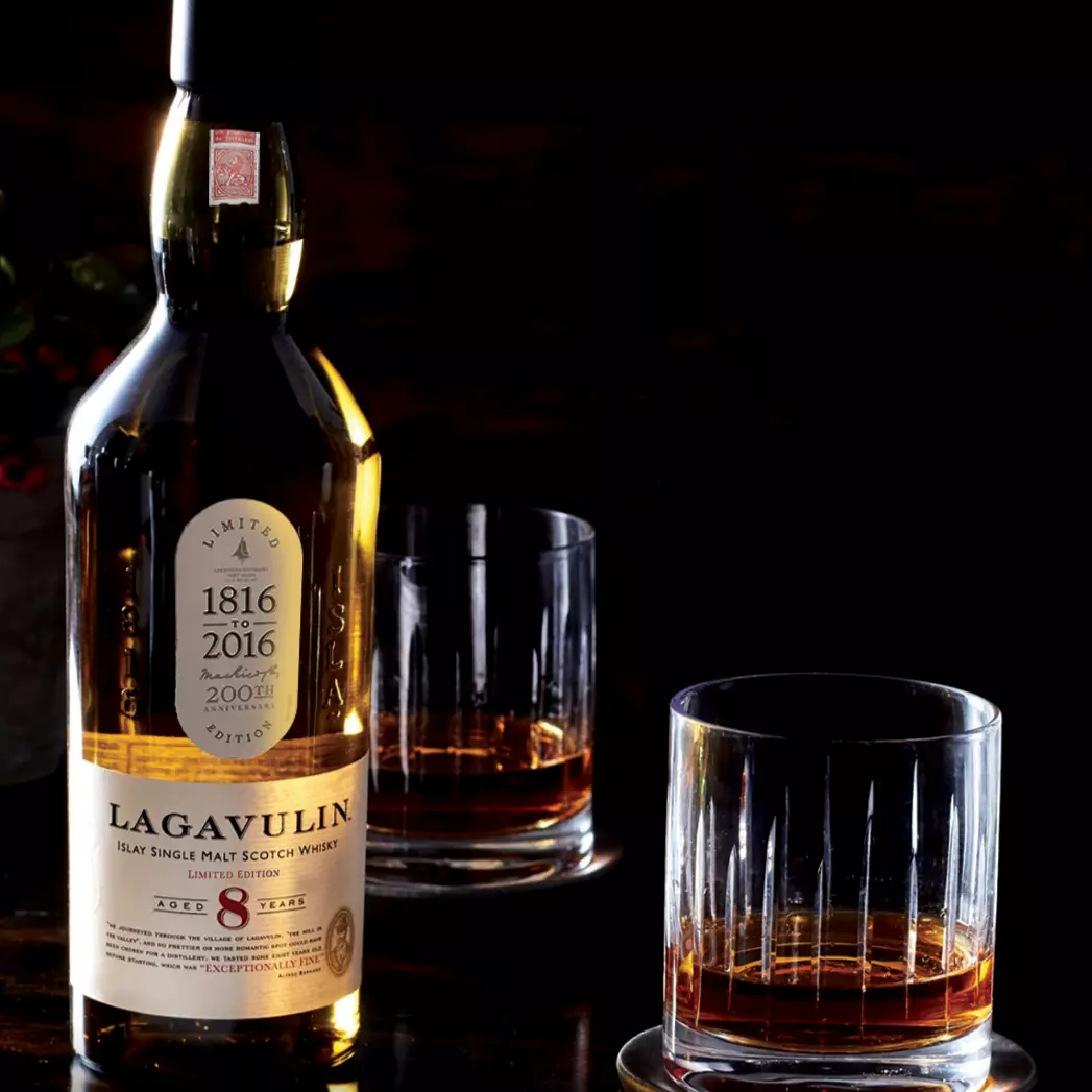 A bottle of Lagavulin 8 Year Old single malt Scotch whisky stands next to two glasses filled with whisky on a dark surface.