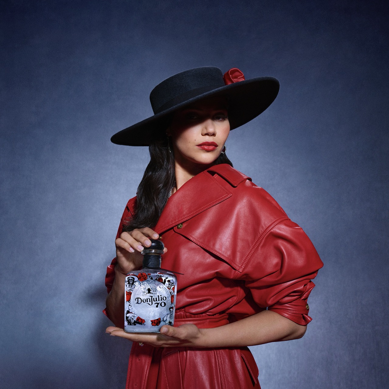 A woman in a red leather coat and wide-brimmed black hat holds a bottle of Don Julio 70 tequila against a dark blue background.