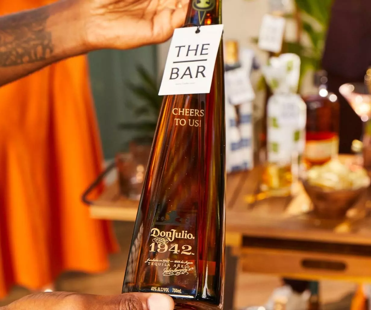 A hand holding a Don Julio 1942 tequila bottle with a tag reading THE BAR Cheers to us! in front of a bar cart with various bottles and barware.