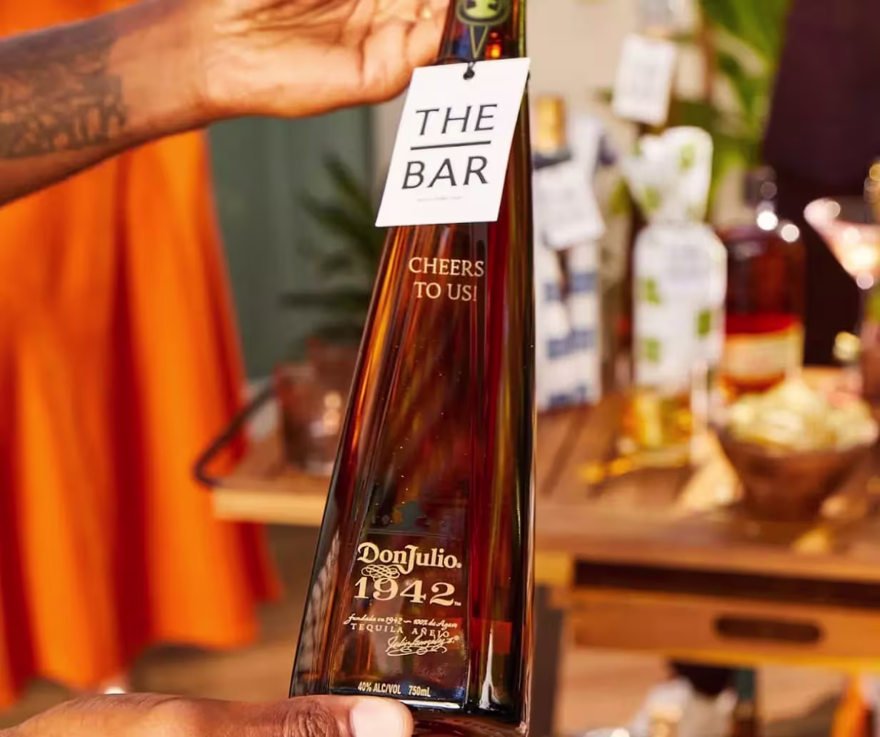 A hand holding a Don Julio 1942 tequila bottle with a tag reading THE BAR Cheers to us! in front of a bar cart with various bottles and barware.