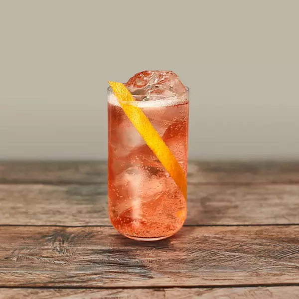 A tall glass filled with an orange-colored iced beverage, garnished with a long twist of orange peel, sits on a wooden surface.