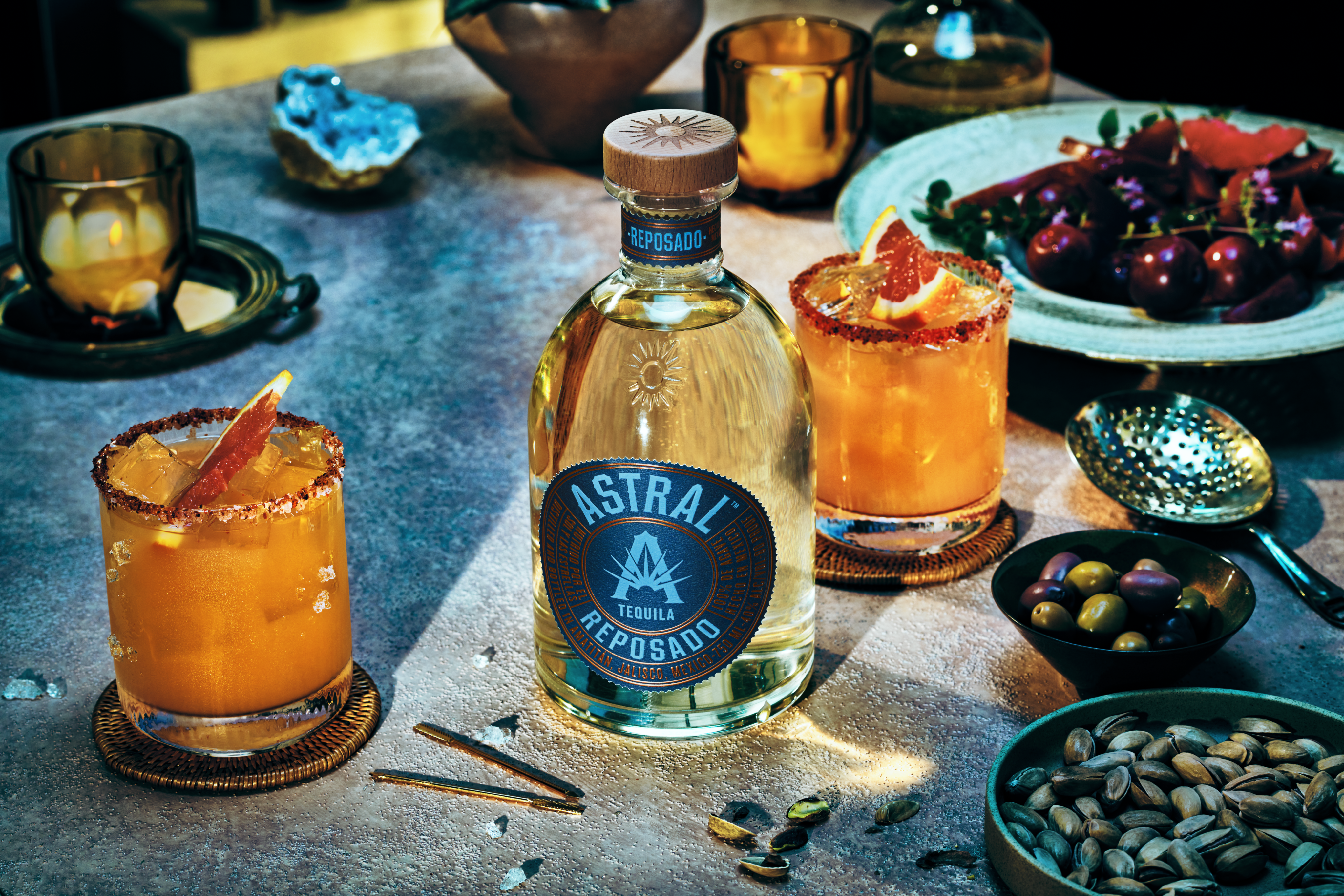 A bottle of Astral Reposado tequila is placed on a table with two cocktails, garnished with orange slices, surrounded by bowls of snacks and a plate of fruit.