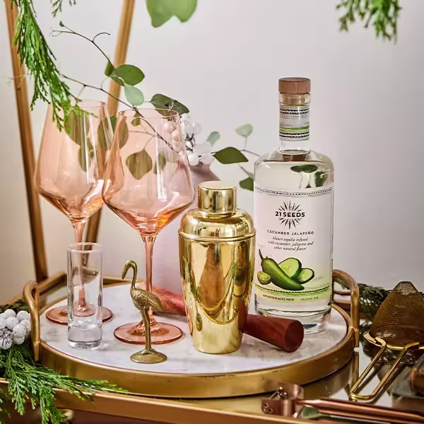 A bar tray with pink wine glasses, a gold cocktail shaker, a shot glass, a bottle of 21 Seeds Cucumber Jalapeño tequila, and decorative greenery.
