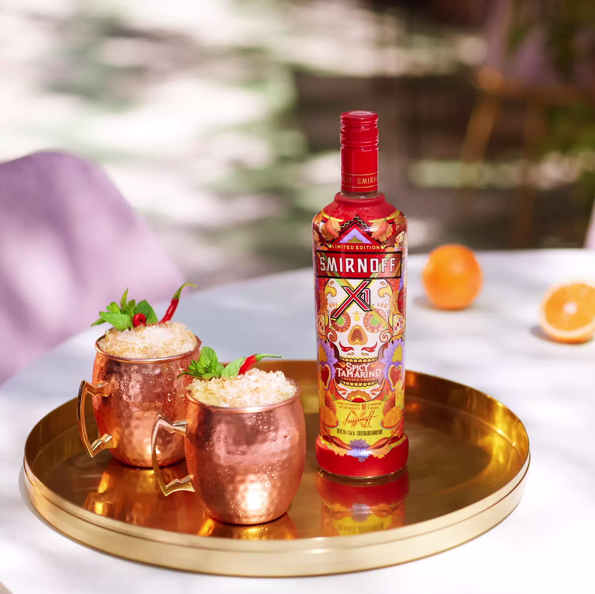 A bottle of Smirnoff Tamarind vodka stands on a gold tray next to two copper mugs filled with ice and garnished with red chili peppers and mint.