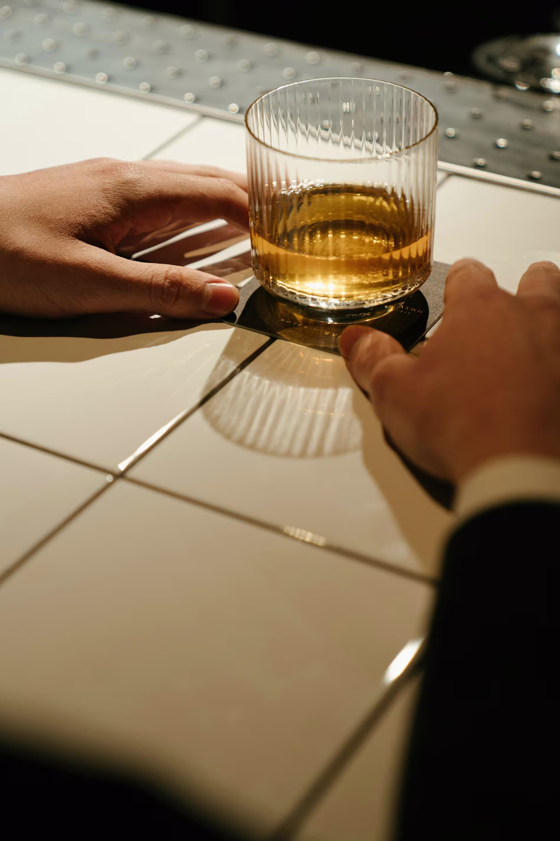 A person in a suit holds a glass of whiskey on a white tiled bar counter.
