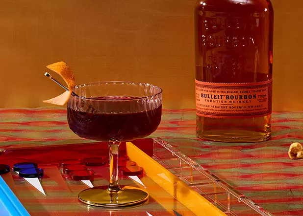 A glass of dark cocktail with an orange peel garnish sits next to a bottle of Bulleit Bourbon on a plaid surface, with colorful backgammon pieces visible nearby.