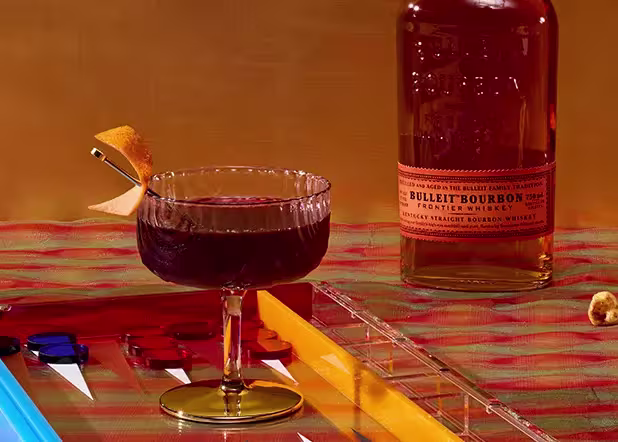 A glass of dark cocktail with an orange peel garnish sits next to a bottle of Bulleit Bourbon on a plaid surface, with colorful backgammon pieces visible nearby.