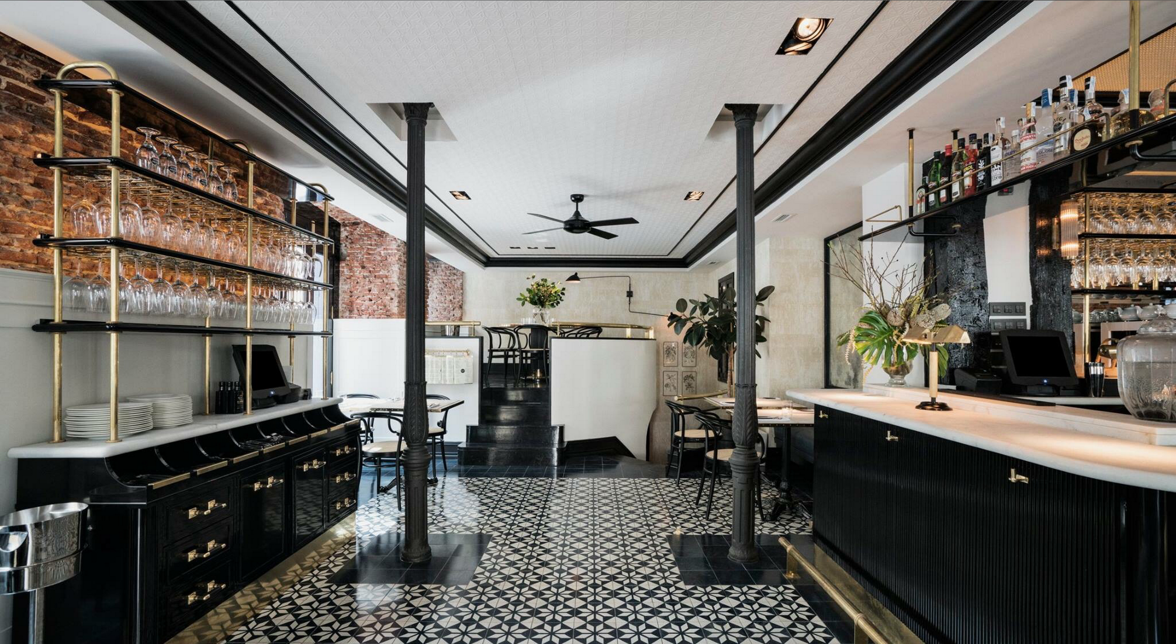 La imagen muestra el interior del restaurante Cannibal Raw. El suelo tiene baldosas geométricas en blanco y negro, y hay un mostrador negro con tiradores dorados en los armarios. Encima del mostrador, hay estantes de vidrio llenos de copas.
