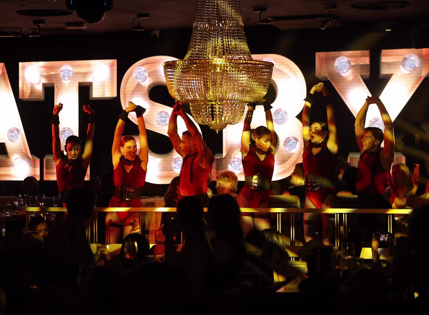 Gatsby: Un espectáculo vibrante y lleno de energía en un entorno glamuroso. Bailarines en atuendos rojos deslumbran bajo la luz de un enorme candelabro, creando una atmósfera sofisticada y teatral.