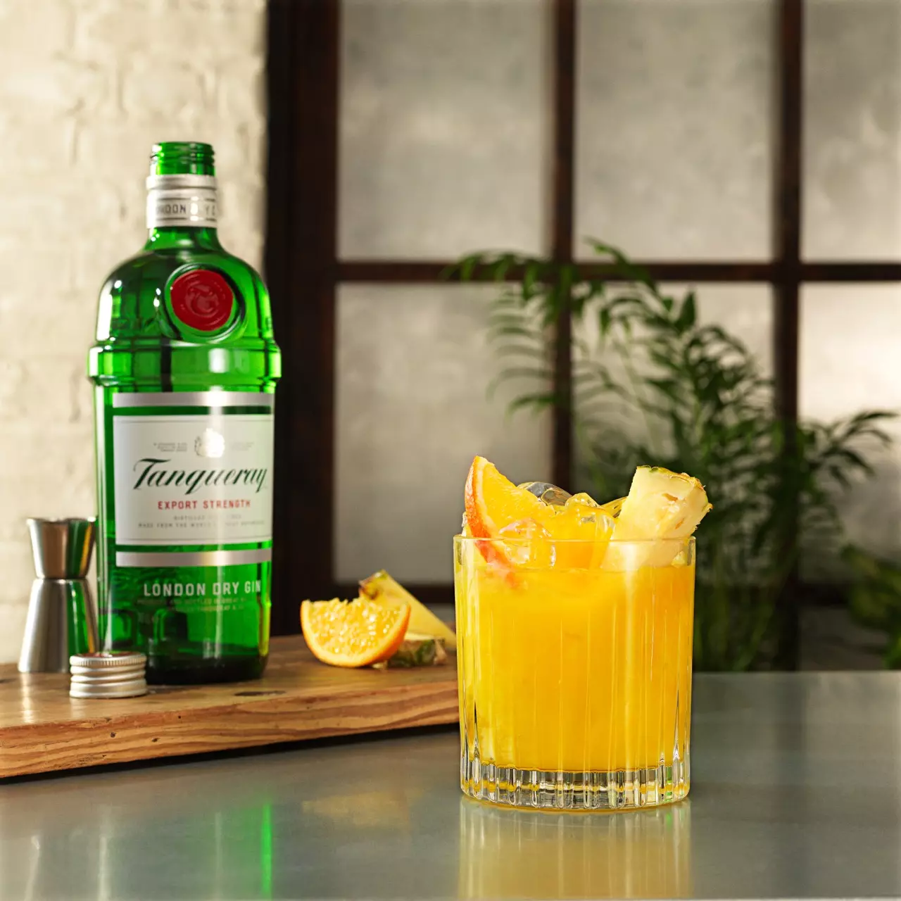 A glass of orange cocktail with ice and fruit garnishes sits on a counter beside a bottle of Tanqueray gin, a jigger, and sliced citrus on a wooden board.