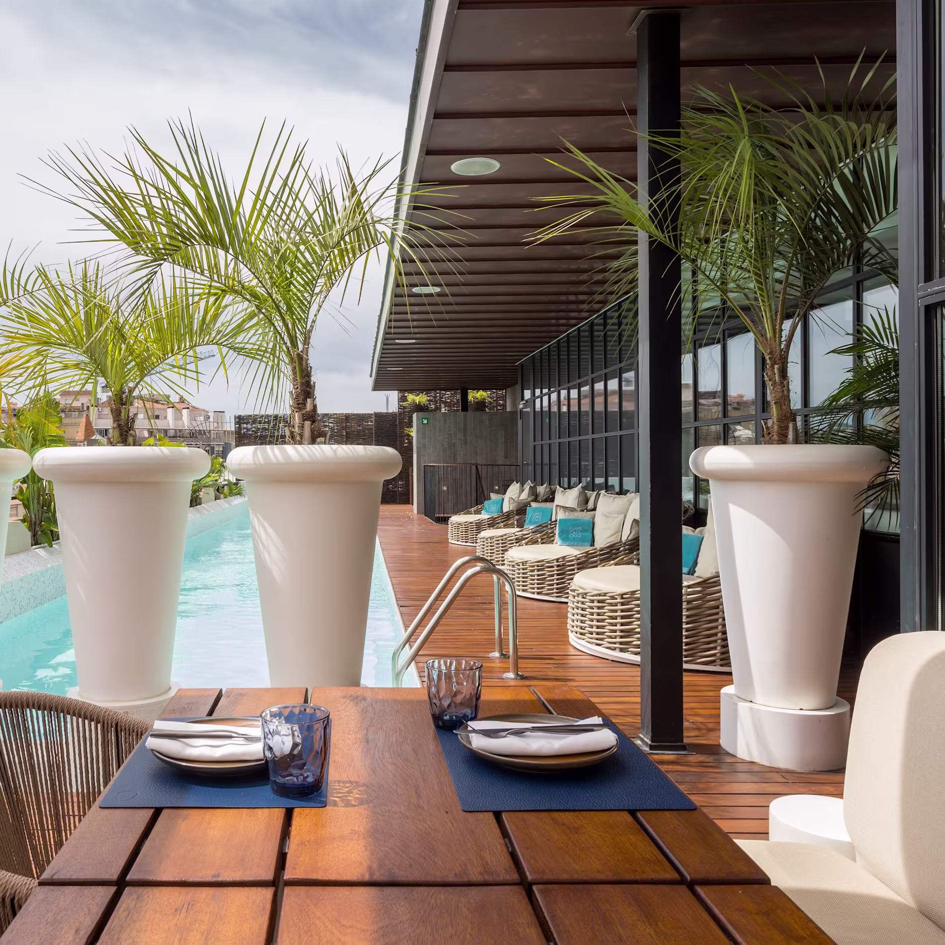 La foto muestra un área de exterior del Rooftop bervena al aire libre con una mesa para dos, con vista a una piscina rodeada de palmeras.