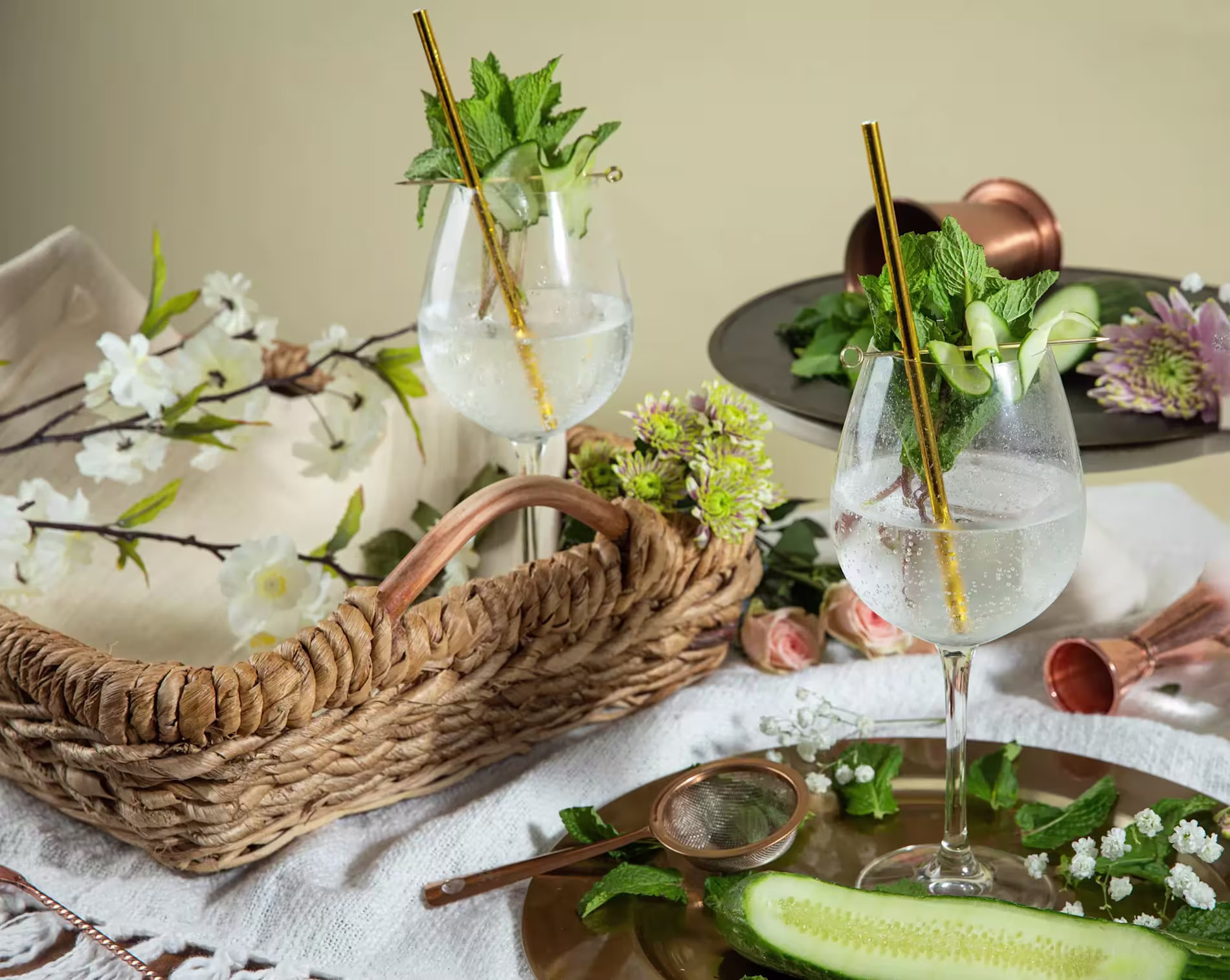 Two gin and tonic cocktails with gold straws, garnished with mint and cucumber, sit on a table with flowers, a wicker basket, and a plate with a cucumber slice.
