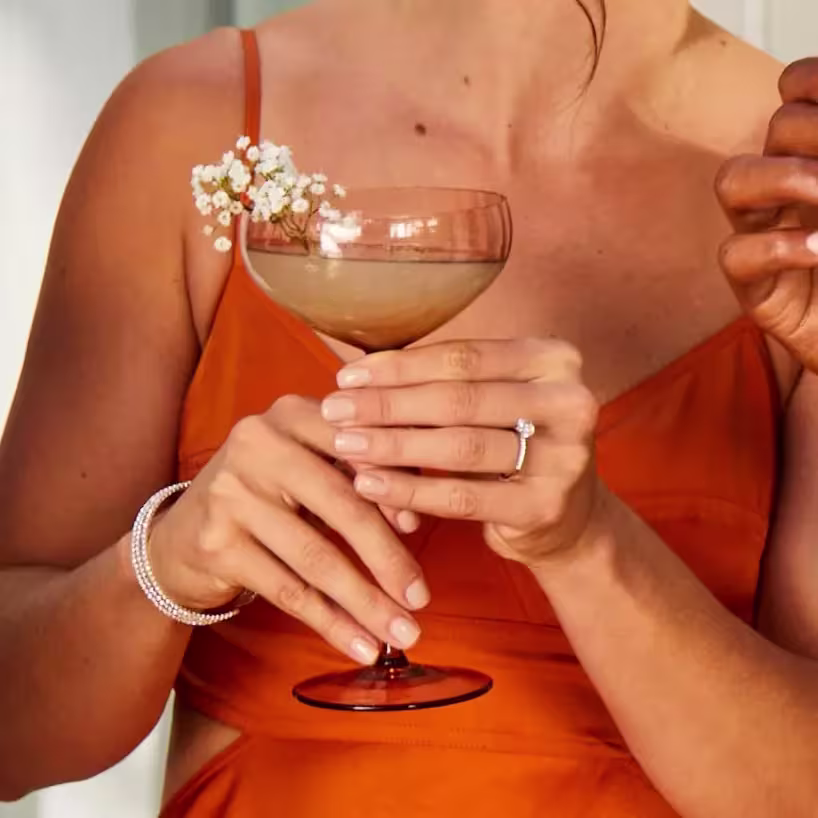 A woman in an orange dress holds a cocktail garnished with small white flowers, wearing a bracelet and an engagement ring.