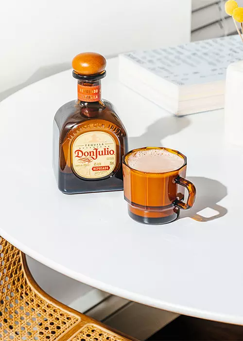 A bottle of Don Julio Añejo tequila and an amber glass mug are placed on a white round table next to a book.