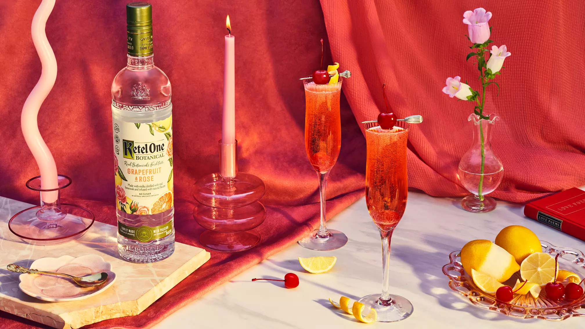 A bottle of Ketel One Botanical Grapefruit & Rose, two pink cocktails with cherries, lemons, pink candles, flowers, and a red matchbox arranged on a marble surface with a pink backdrop.