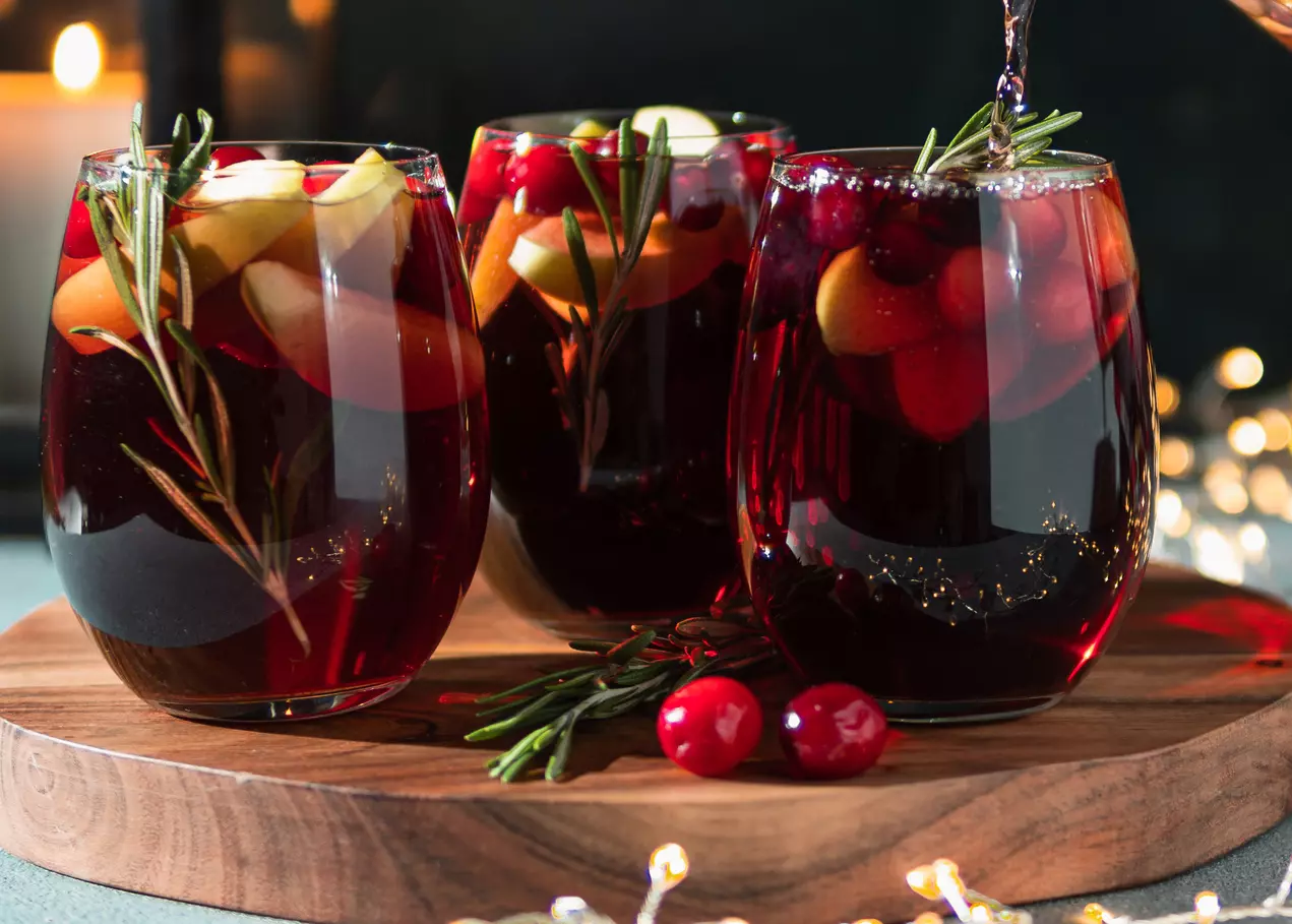 Three glasses of red sangria with apple slices, cranberries, and rosemary sprigs on a wooden tray, with a festive background.