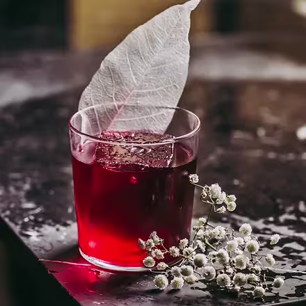 En Farola, un cóctel rojo intenso con una hoja blanca decorativa y flores delicadas crea una experiencia visual y gustativa única.