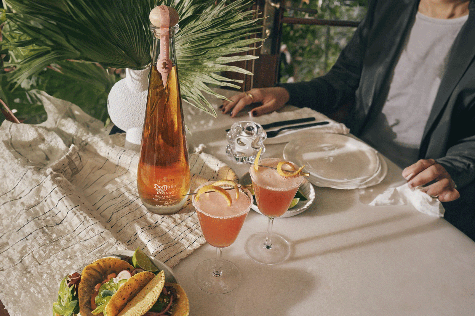 A table set with a bottle of tequila, two pink cocktails with orange peels, a plate of tacos, a vase with greenery, and a person sitting at the table.