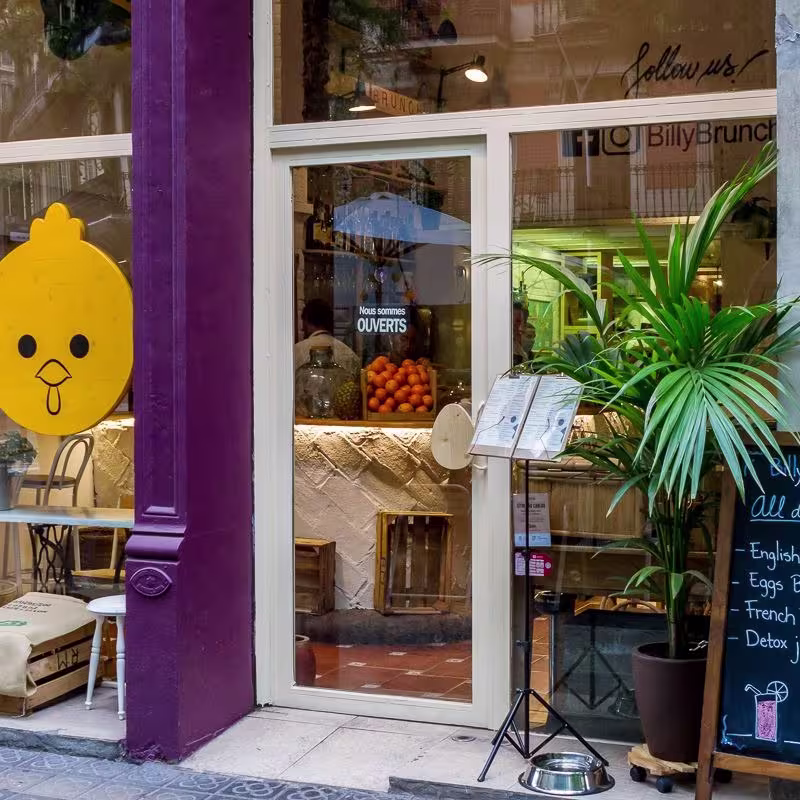 Fachada de Billy Brunch, acogedor café en Barcelona con cartel que anuncia desayunos todo el día como Eggs Benedict, French Toast y jugos detox.