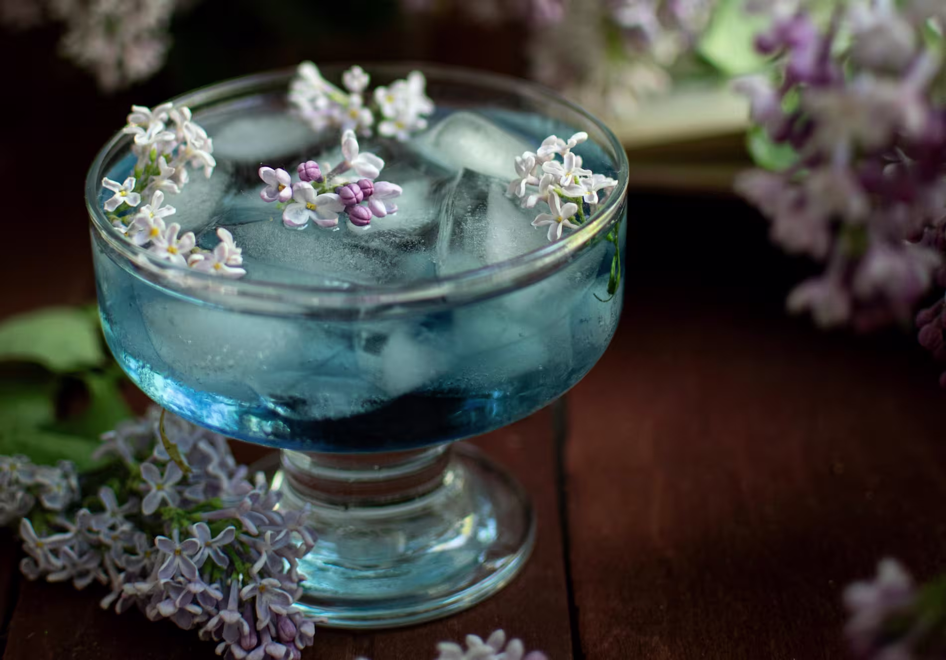 A glass cup filled with blue iced beverage, garnished with small lilac flowers, sits on a wooden surface surrounded by more lilac blooms.