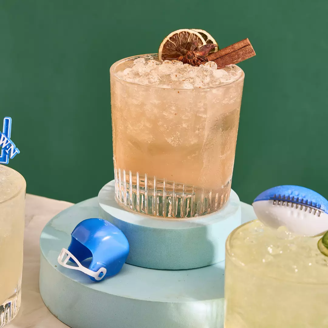 A cocktail garnished with a dried citrus slice and cinnamon stick sits on a circular pedestal, next to a small blue toy football helmet and a toy football.