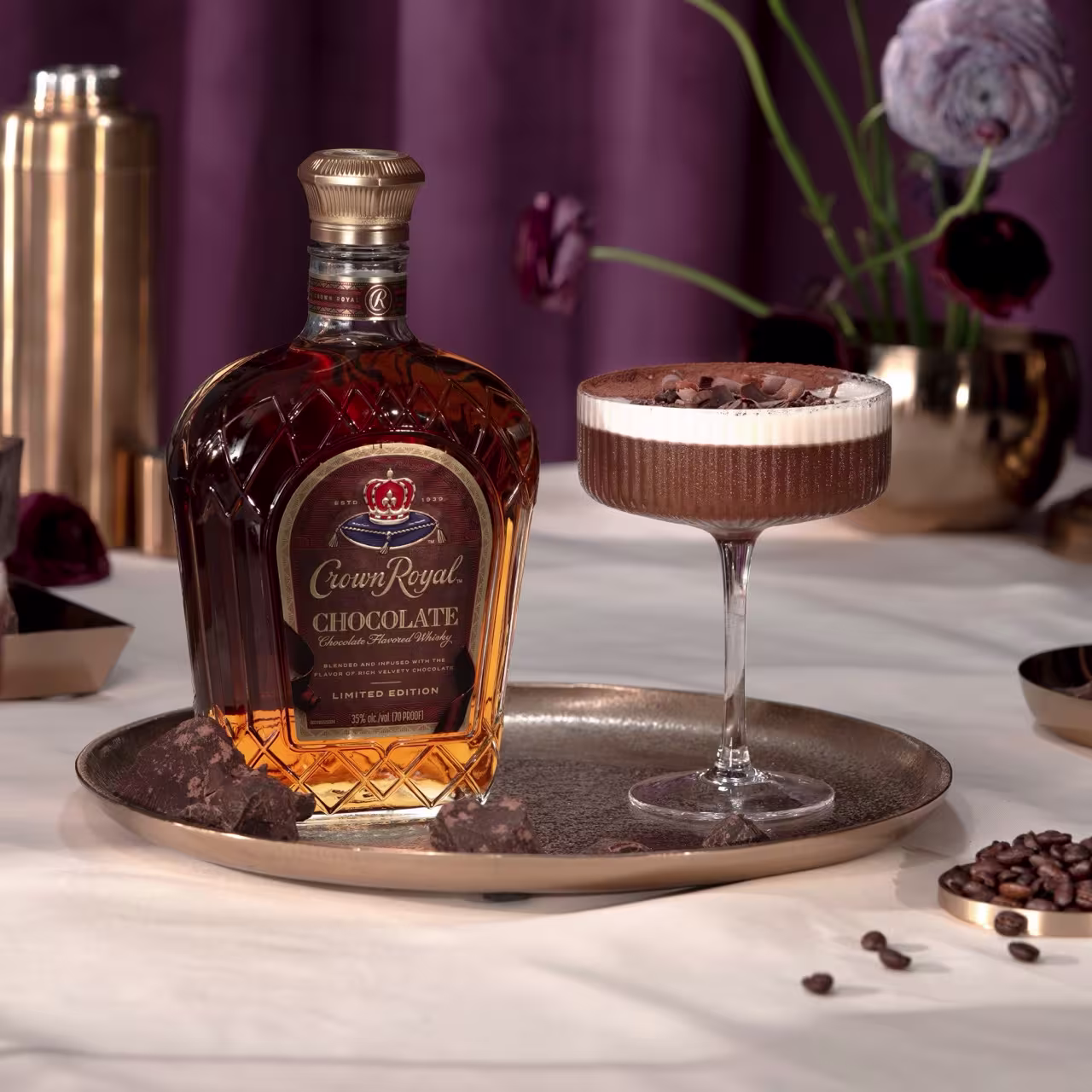 A bottle of Crown Royal Chocolate whiskey and a chocolate cocktail on a gold tray, with chocolate pieces and coffee beans nearby.