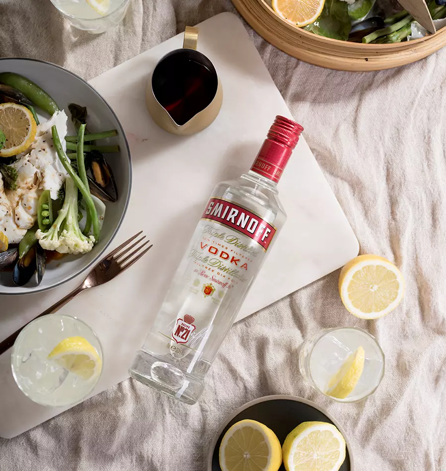 A bottle of Smirnoff vodka on a white surface, surrounded by plates of food, lemon slices, drinks, and a small jug of dark liquid.