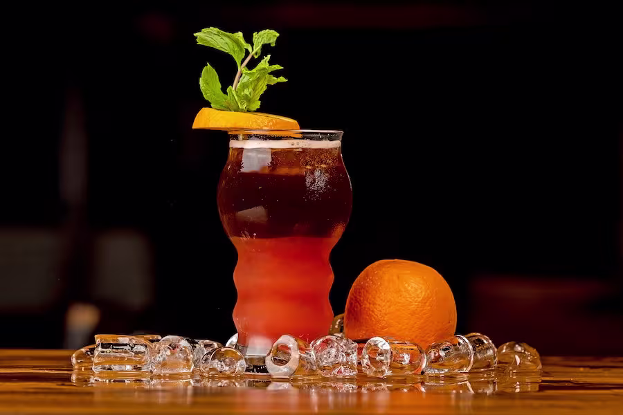 A cocktail garnished with an orange slice and mint is surrounded by ice cubes and a whole orange on a wooden surface with a dark background.