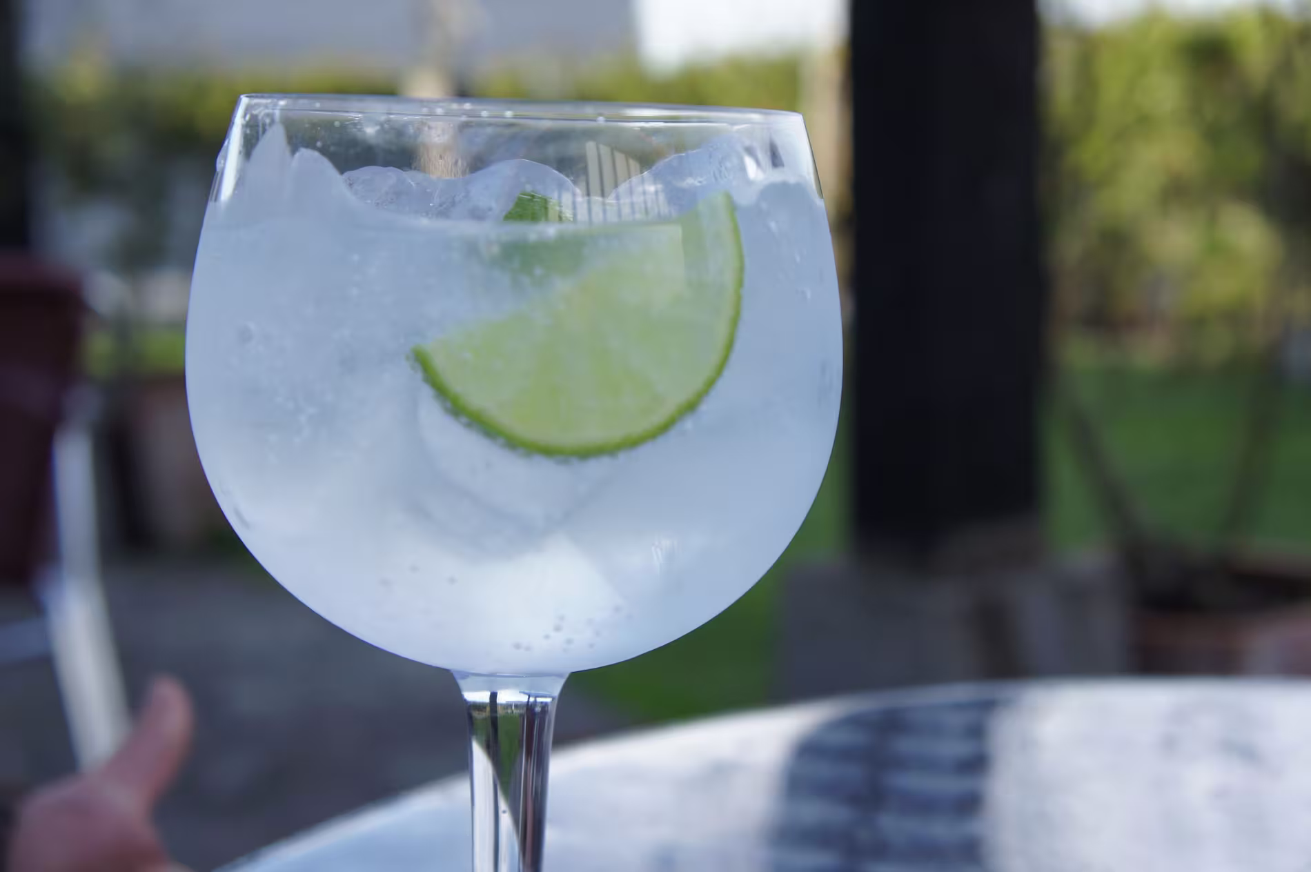 A clear glass filled with ice, fizzy liquid, and a slice of lime sits on a reflective outdoor table with greenery in the background.
