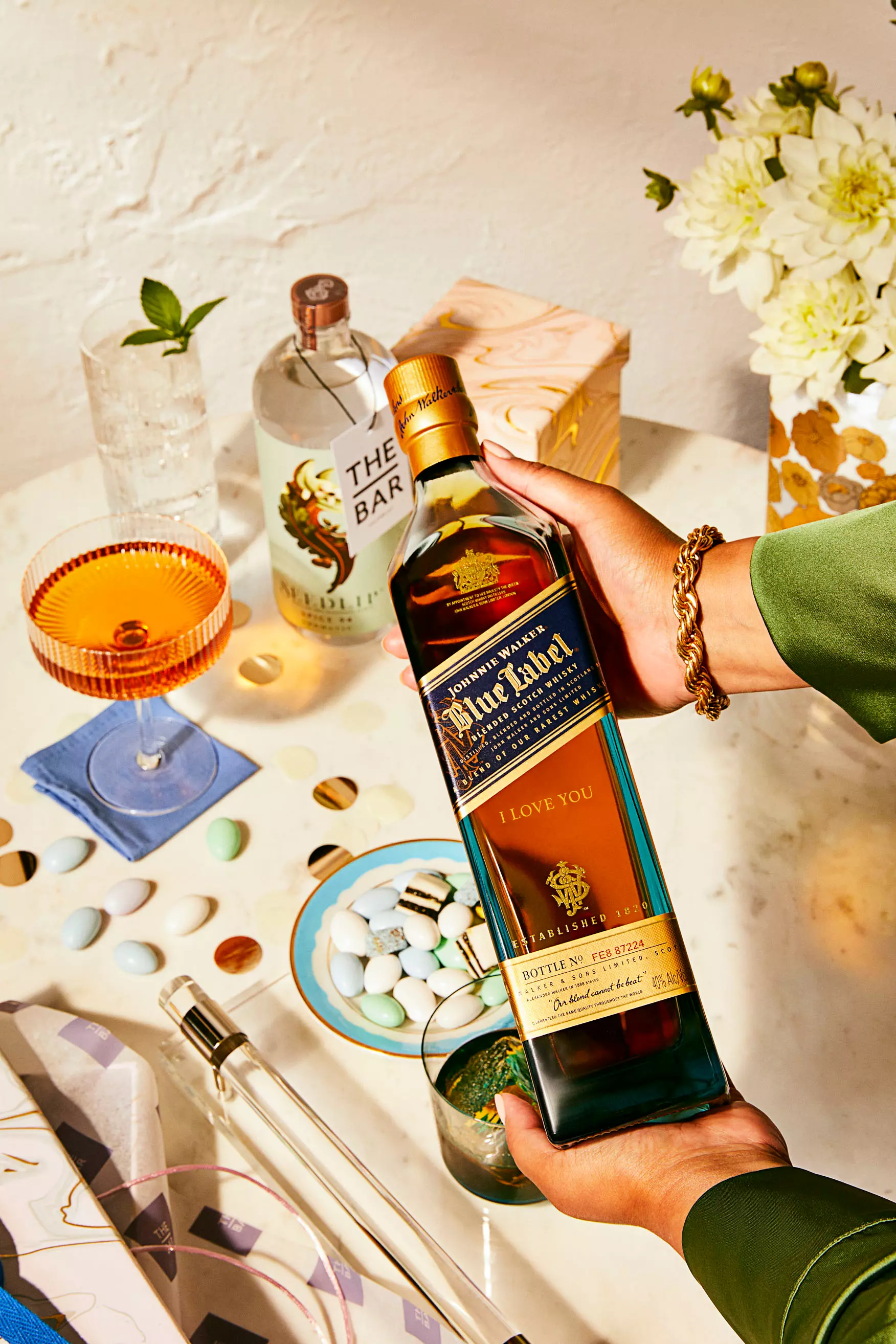 A person holds a bottle of Johnnie Walker Blue Label whisky labeled I Love You over a table set with drinks, flowers, and snacks.
