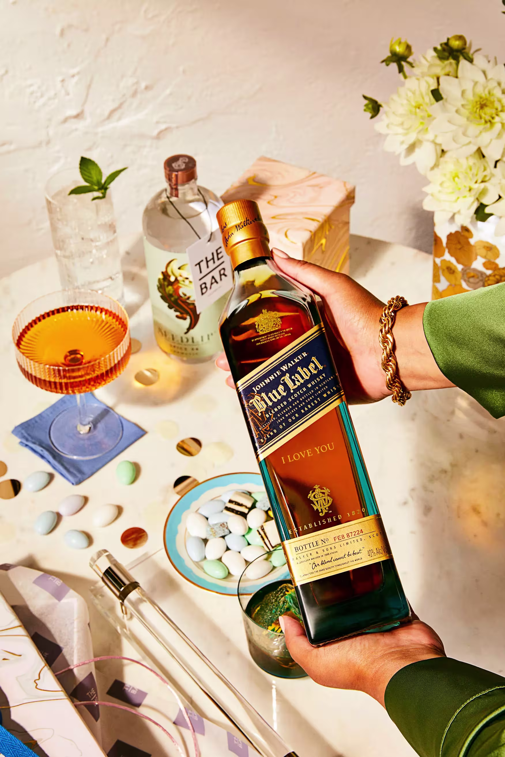 A person holds a bottle of Johnnie Walker Blue Label whisky labeled I Love You over a table set with drinks, flowers, and snacks.