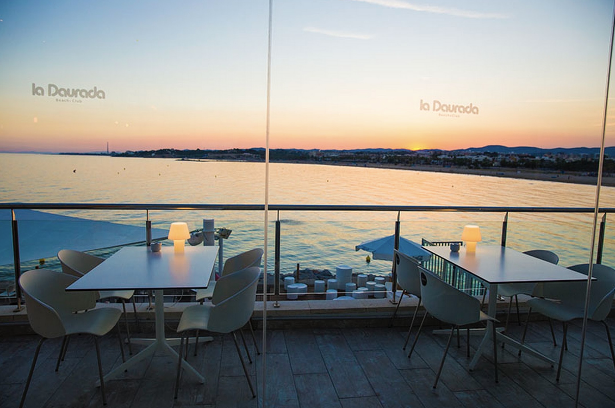 Terraza de La Daurada, con mesas minimalistas, sillas blancas y vistas panorámicas al mar al atardecer, creando un ambiente tranquilo.