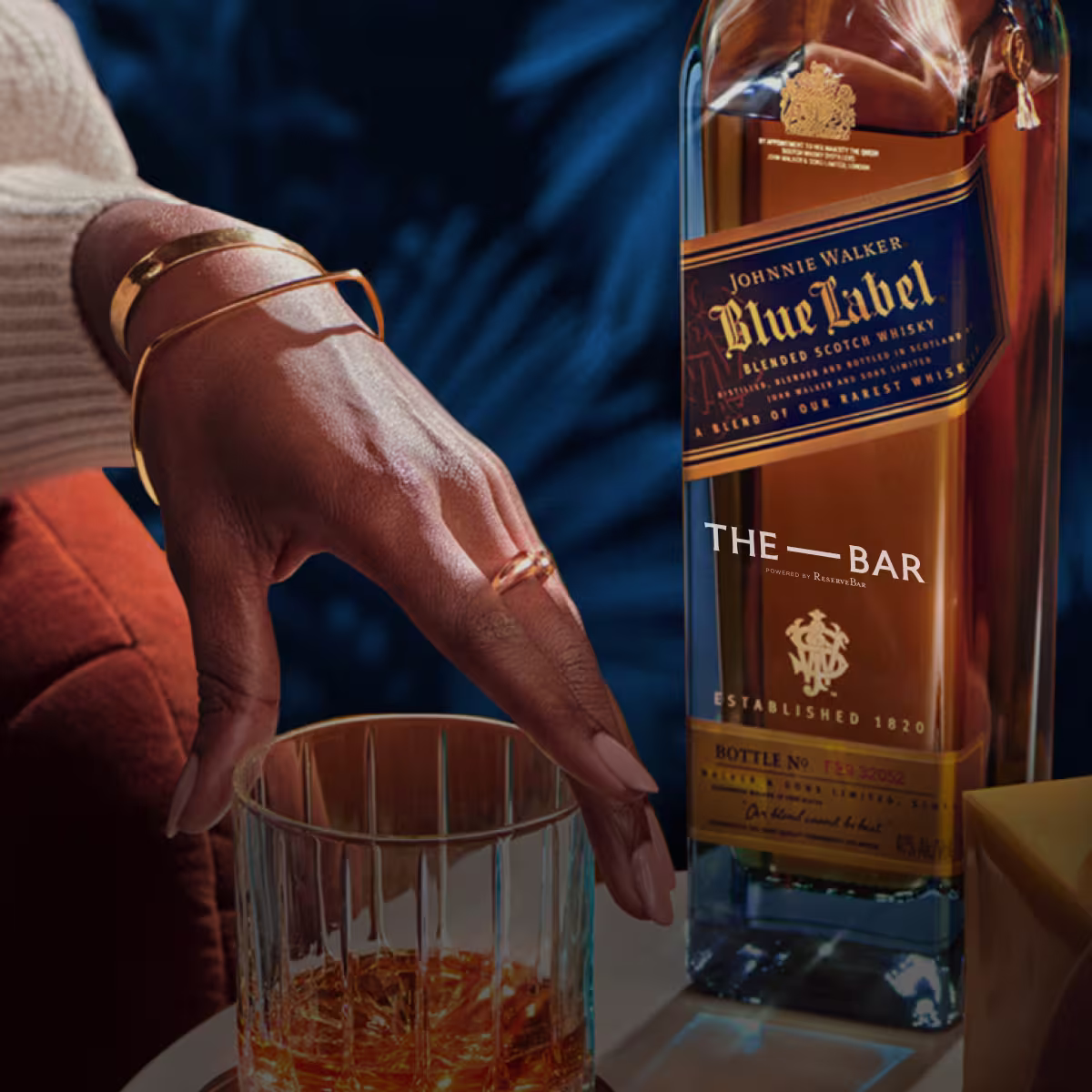 A hand with gold jewelry reaches for a glass of whiskey beside a bottle of Johnnie Walker Blue Label on a table.