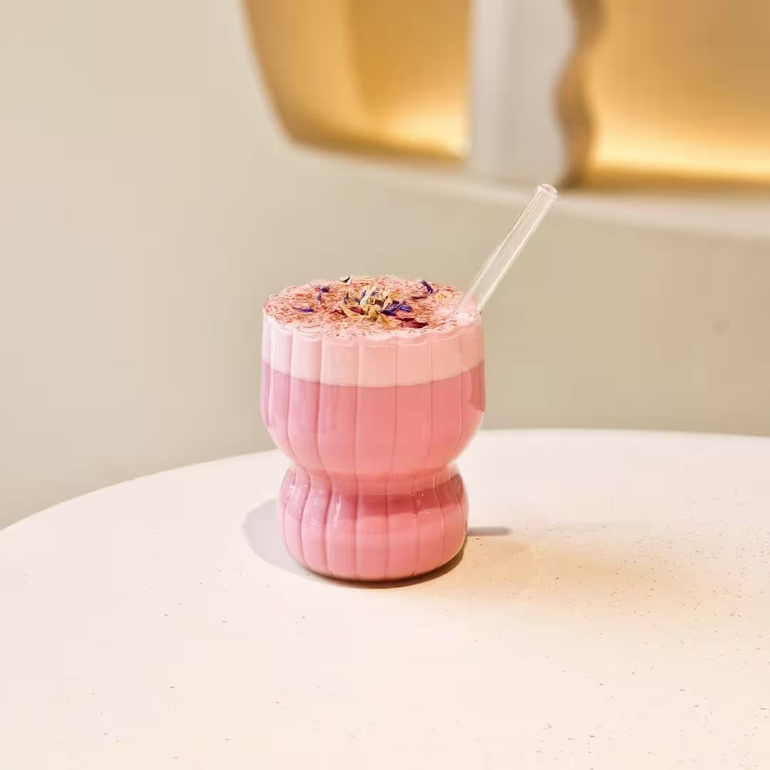 Bebida rosa servida en un vaso curvo a juego con espuma decorada con pétalos y pajita transparente sobre una mesa blanca de estilo minimalista.