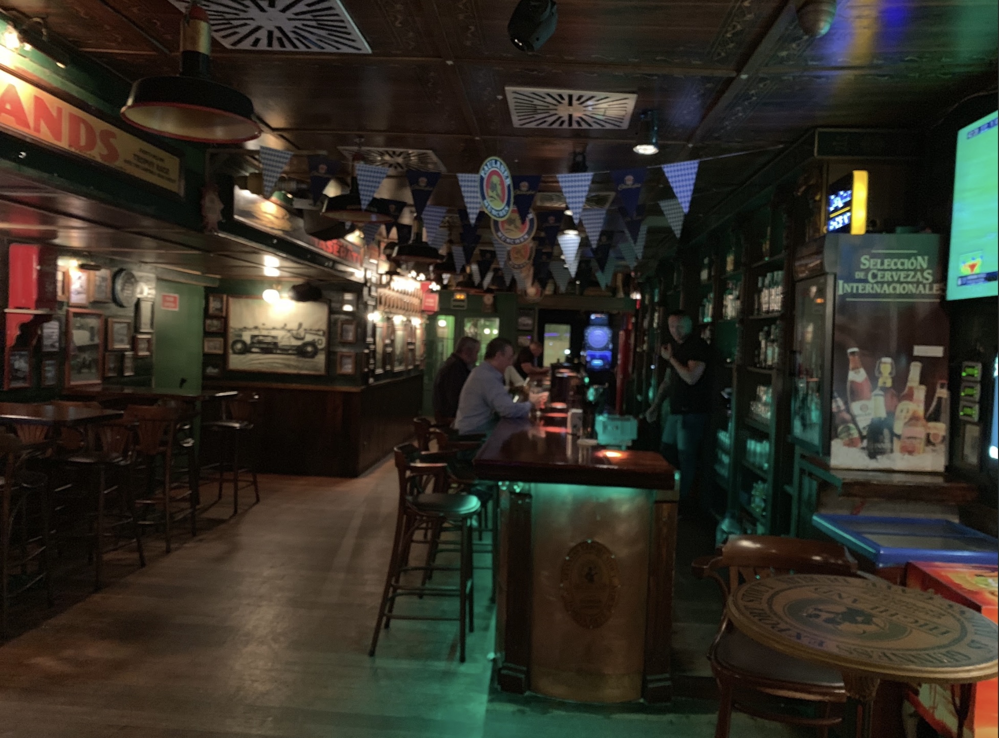 La imagen muestra el pub Higland de estilo clásico con decoración en madera oscura, banderines colgantes y carteles vintage. El ambiente es acogedor y tradicional, con clientes sentados en la barra.