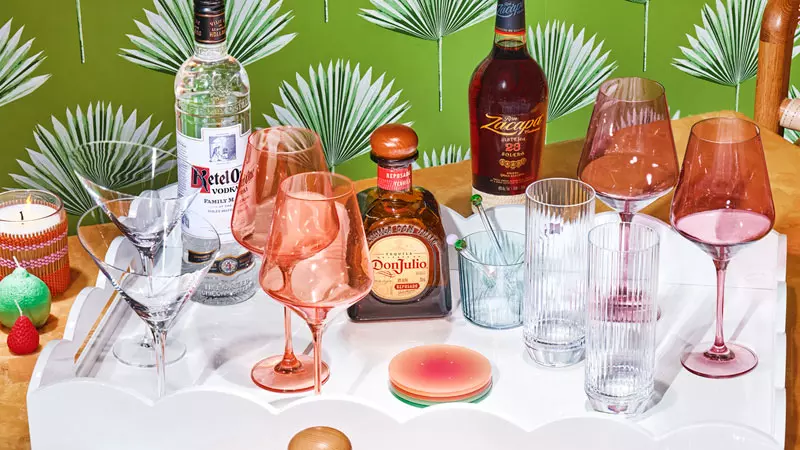 A white tray holding assorted liquor bottles, drinking glasses, and cocktail glasses sits on a table with a green leafy-patterned background.