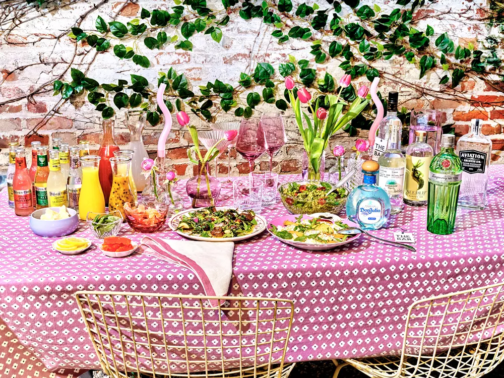 A table set for an outdoor meal with salads, condiments, various drinks, bottles of alcohol, flowers in vases, and glassware, against a brick wall with green ivy.