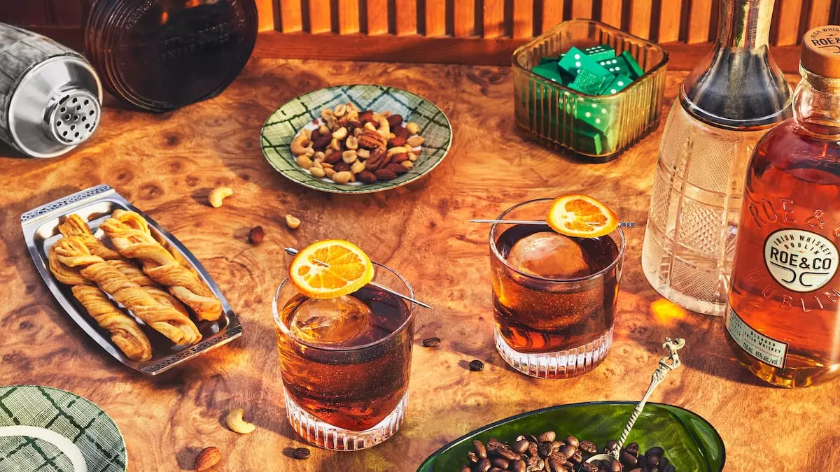 Two glasses of dark cocktails with orange garnish sit on a wooden table among snacks, a cocktail shaker, a bottle of whiskey, and a bowl of dice.