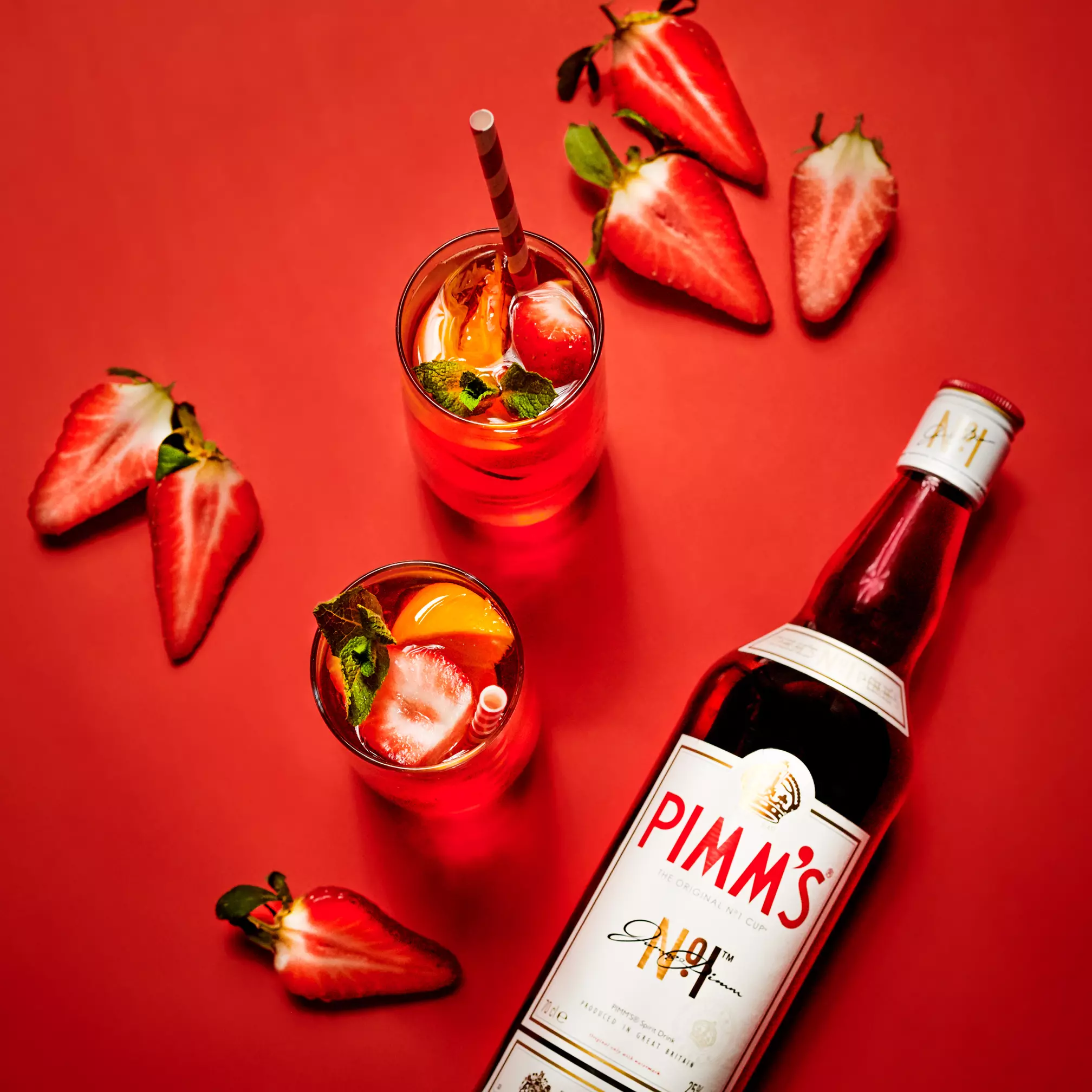 A bottle of Pimms, two glasses of red cocktail with fruit and mint, and several halved strawberries on a red background.
