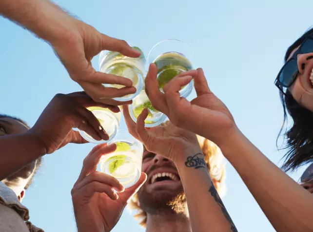 Four people clinking plastic cups with drinks and lime slices outdoors, viewed from below with a clear blue sky in the background.