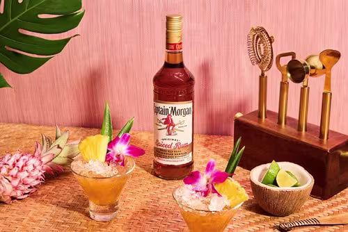 A bottle of Captain Morgan Original Spiced Rum is placed on a woven mat with two tropical cocktails, a pineapple, lime wedges, and a cocktail tool set against a pink background.