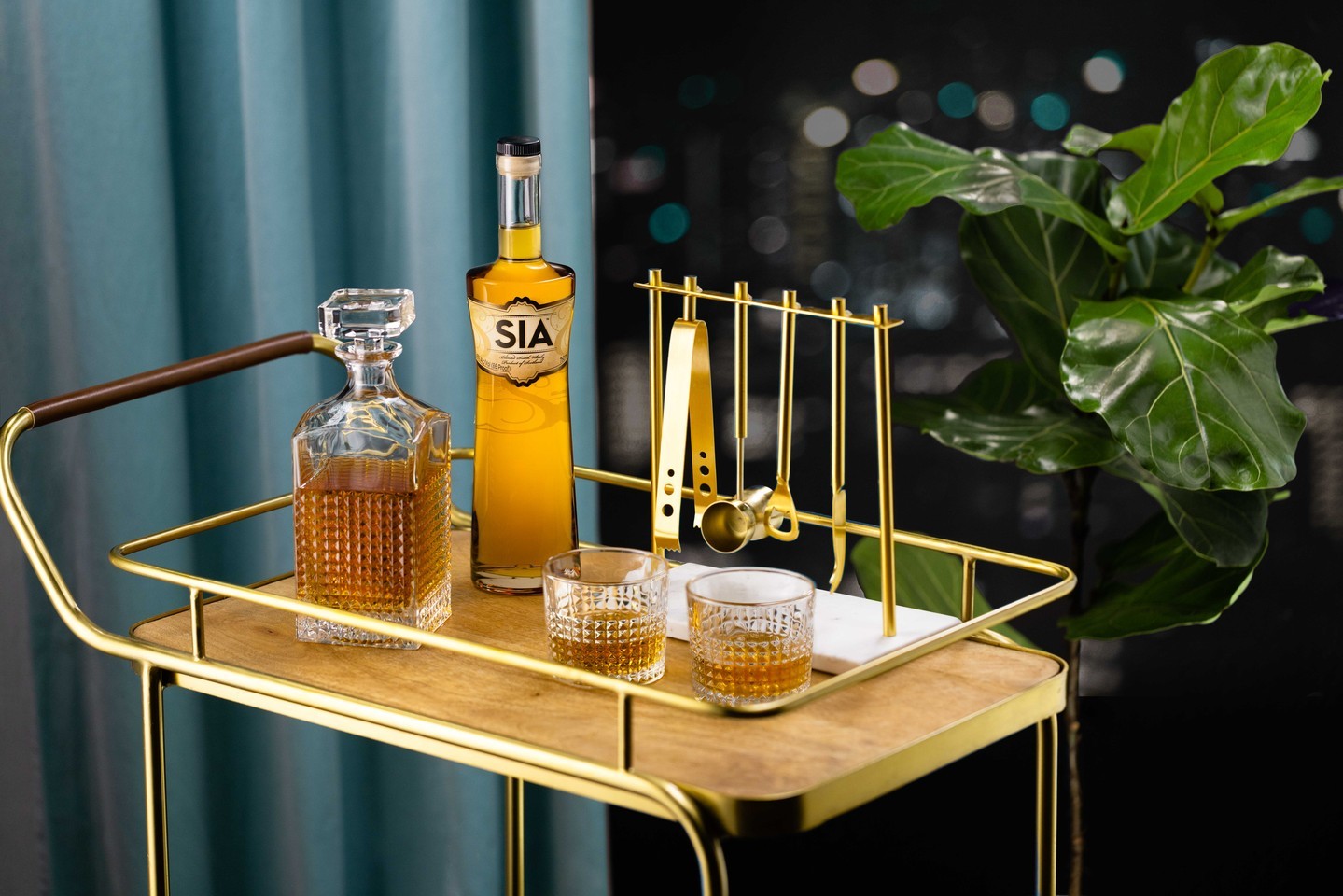 A gold bar cart with a decanter, a bottle of SIA Scotch Whisky, two filled glasses, bar tools, and a potted plant in the background.