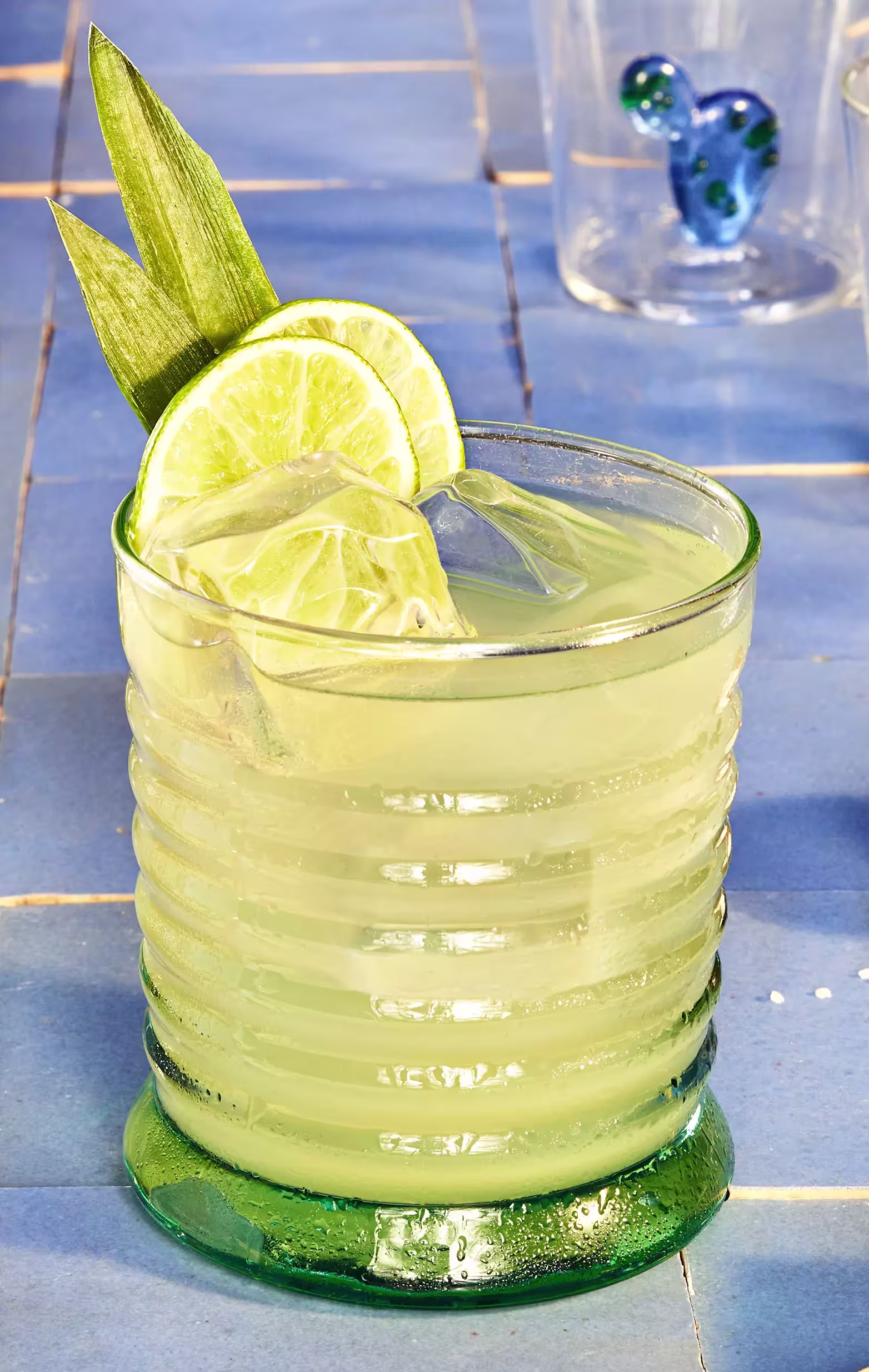 A glass of light green cocktail with ice cubes, lime slices, and two green leaves garnish, placed on a blue tiled surface.