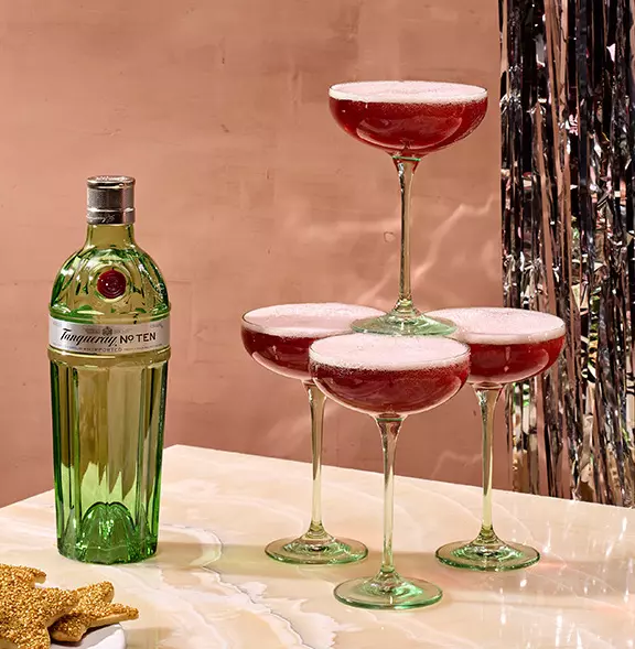 A bottle of Tanqueray No. Ten gin next to four coupe glasses filled with a red cocktail, stacked in a pyramid on a marble surface.