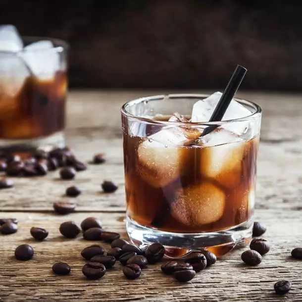A glass filled with iced coffee and a black straw sits on a wooden surface, surrounded by scattered coffee beans. Another similar glass is in the background.
