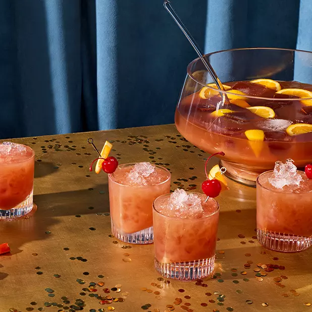 A punch bowl filled with an orange-colored drink and fruit slices sits on a table, surrounded by four glasses of the same drink garnished with cherries and orange slices.