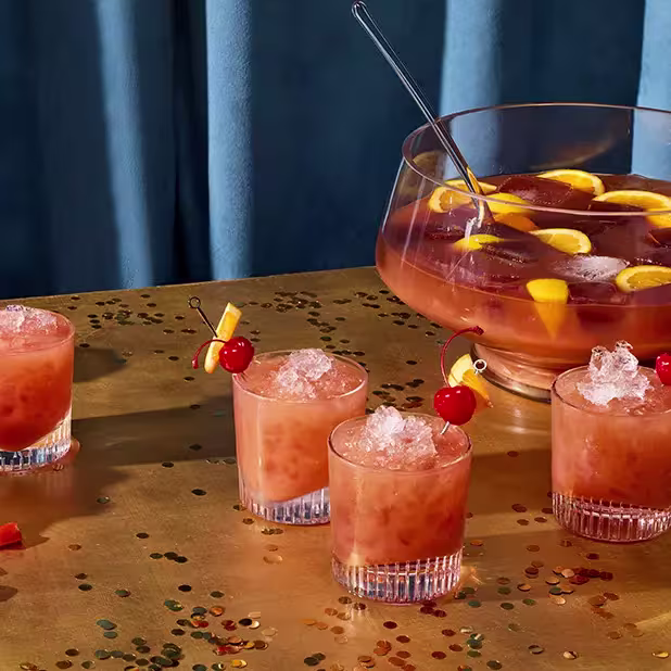 A punch bowl filled with an orange-colored drink and fruit slices sits on a table, surrounded by four glasses of the same drink garnished with cherries and orange slices.