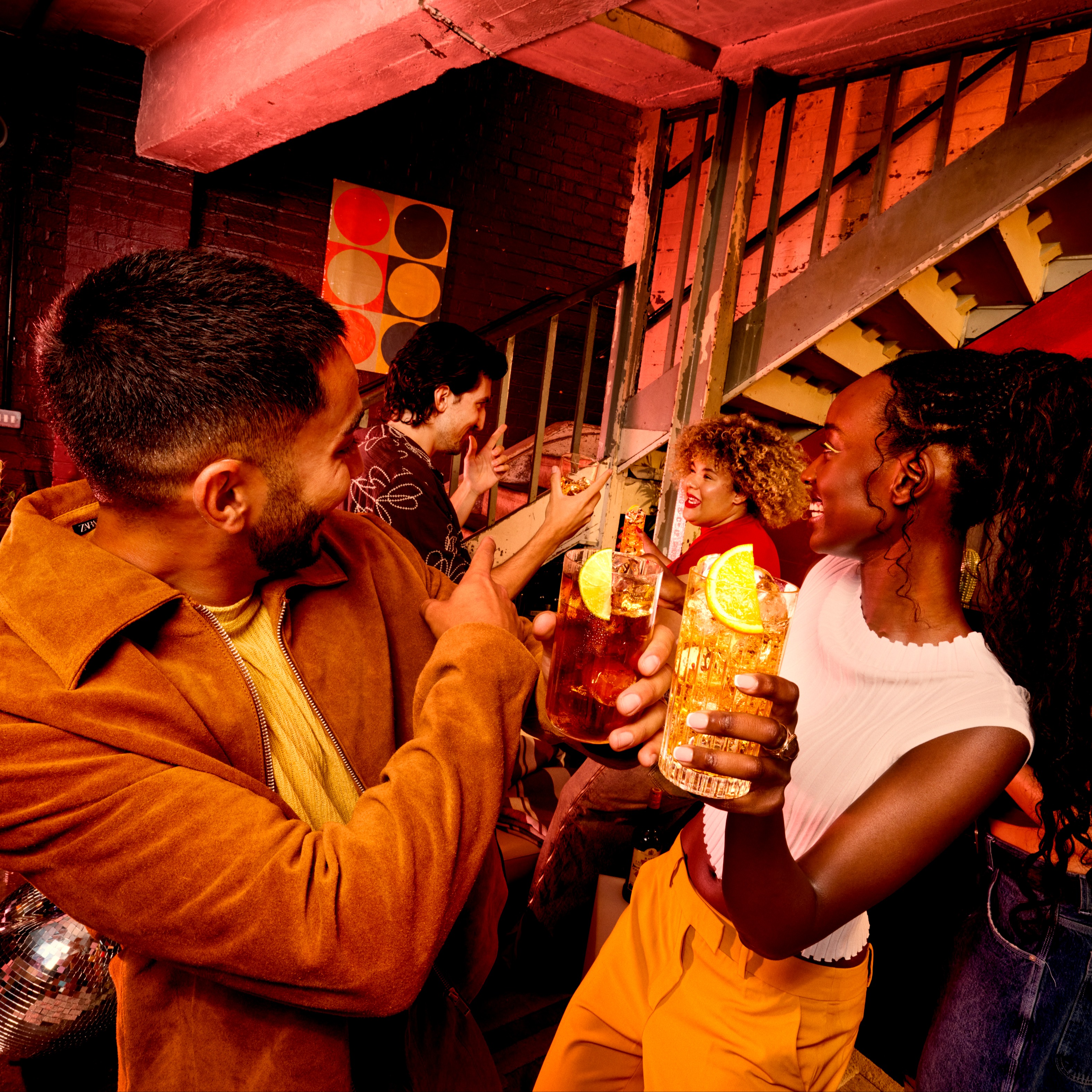 Doi prieteni ciocnesc paharele de cocktail „highball” la o petrecere animată acasă.